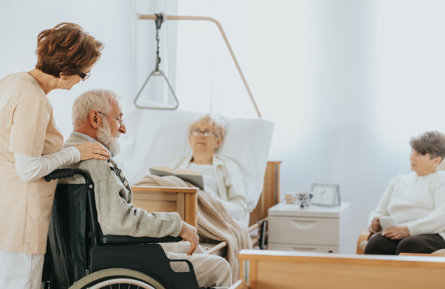 A female caregiver comforting an elderly man in a wheelchair in a bright nursing home room with other residents.