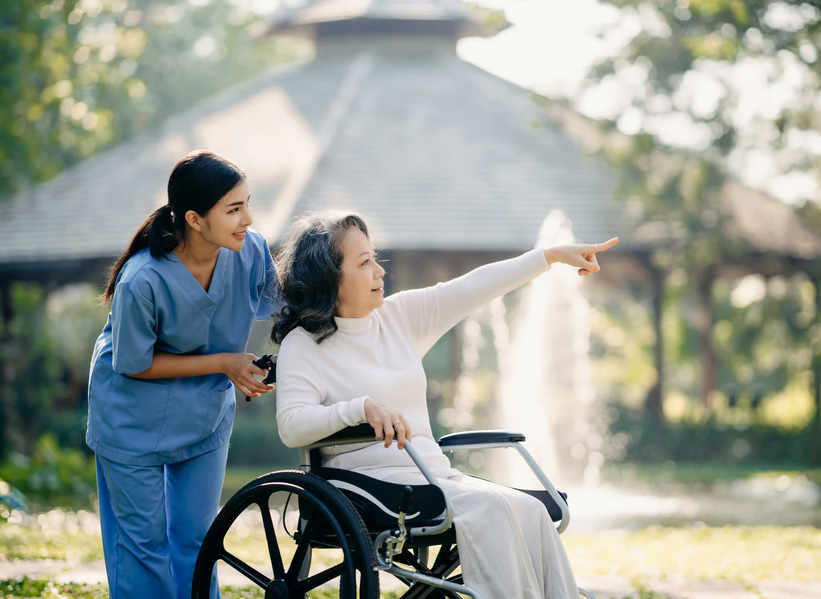 Compassionate young female caregiver in blue medical scrubs assists a smiling elderly Asian woman seated in a wheelchair during an outdoor walk. The elderly woman is pointing towards a sun-dappled fountain and park gazebo.