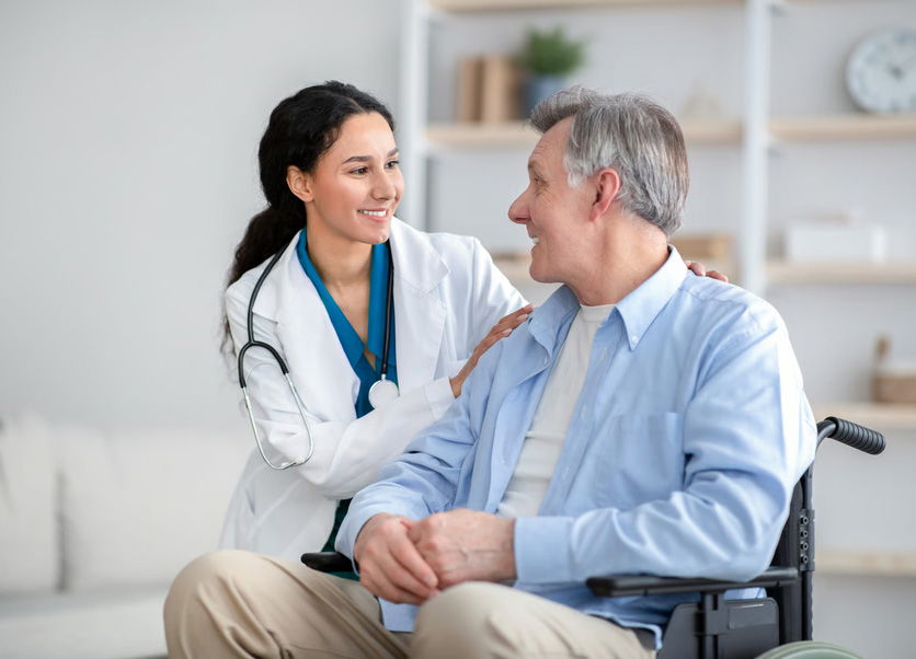 A smiling female doctor in a white lab coat comforting a senior man in a wheelchair.