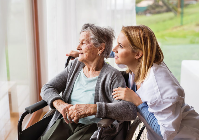 Female caregiver comforting an elderly woman in a wheelchair looking out a window.