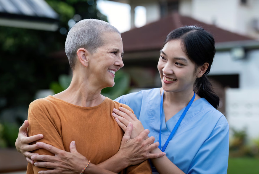 A female nurse in blue scrubs smiling and embracing a female cancer survivor with short hair outdoors.