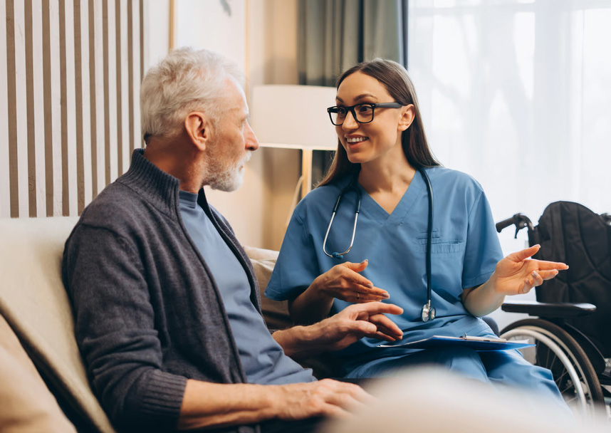 Female home health nurse in blue scrubs talking with a senior man on a couch.