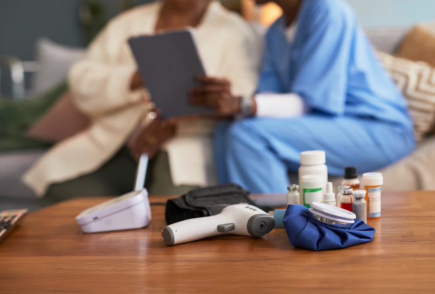 Medical monitoring tools on a table with a blurred nurse and senior patient using a digital tablet in the background.