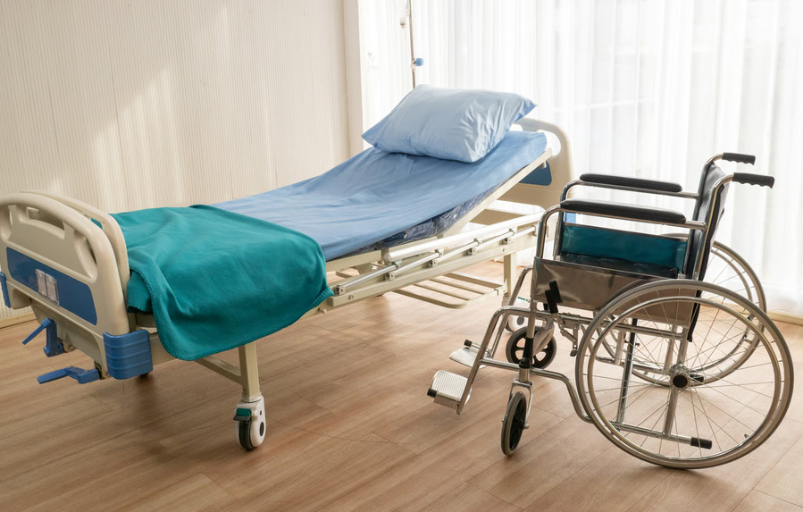 An adjustable medical hospital bed with blue sheets next to a manual wheelchair in a bright room.