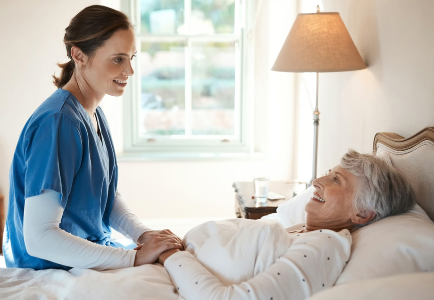 A compassionate hospice nurse holding the hand of a smiling senior woman resting in bed during an in-home care visit.