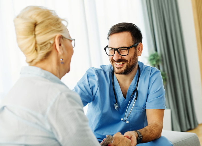 Smiling male nurse in blue scrubs holding hands with an elderly woman during a home care visit.