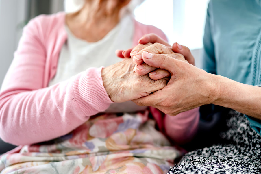 A caregiver gently holding the hands of an elderly woman wearing a pink cardigan, showing support and senior care.