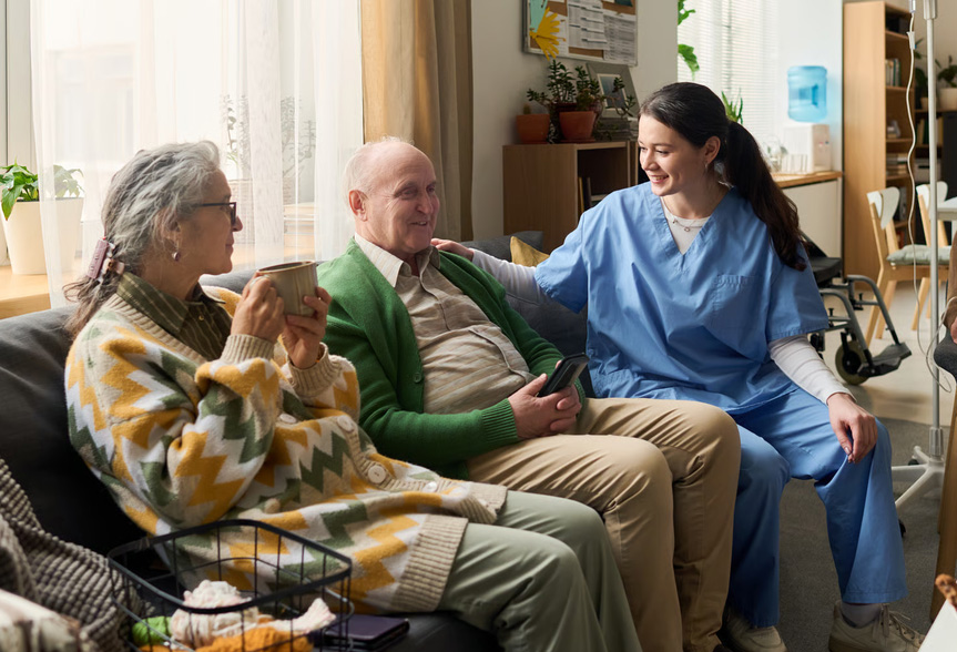 A female nurse in blue scrubs talking and smiling with an elderly couple sitting on a couch.