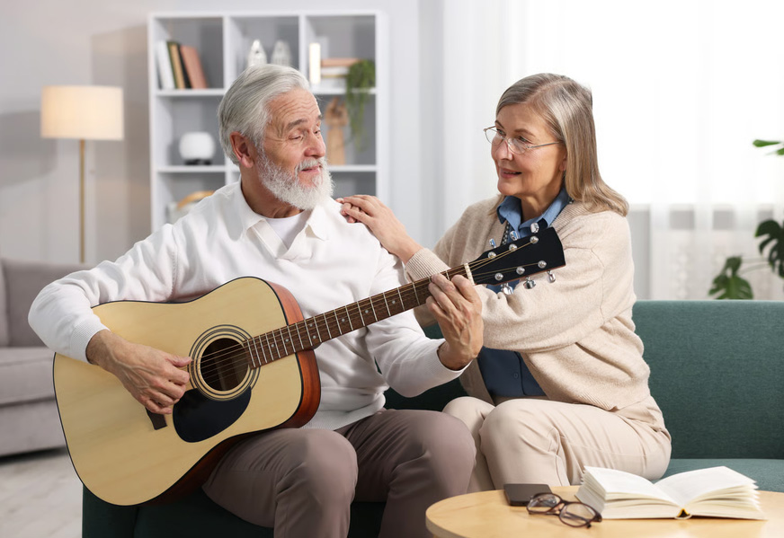 Elderly couple participating in music therapy with the husband playing a guitar on a sofa.