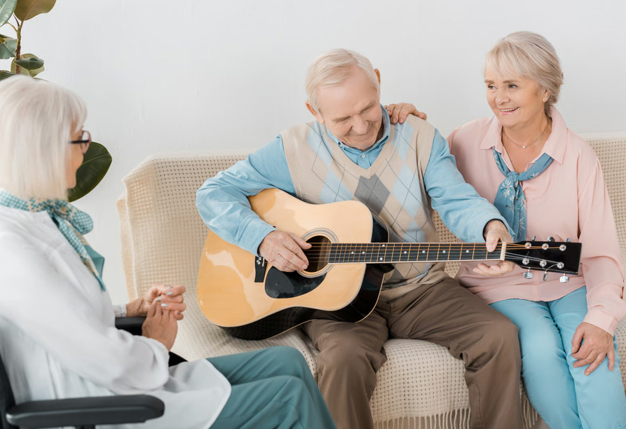 Senior residents gathered on a couch enjoying music therapy with a man playing acoustic guitar.