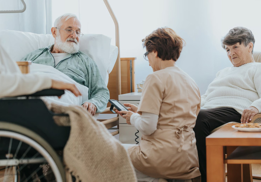 A female caregiver checking a device next to an elderly man in a hospital bed and a senior woman sitting nearby.